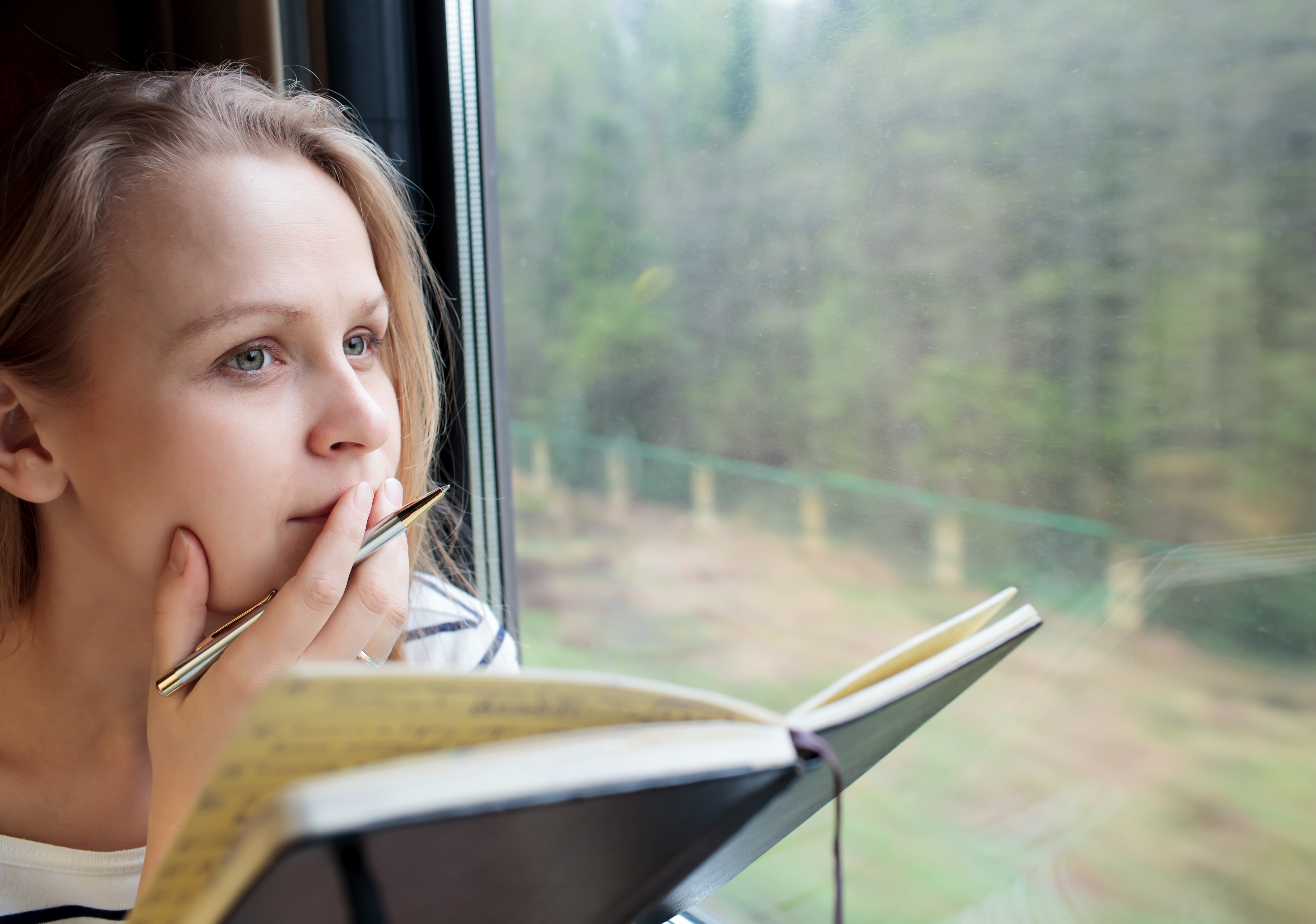 Young woman on a train writing notes in a diary or journal staring thoughtfully out of the window with her pen to her lips as she thinks of what to write